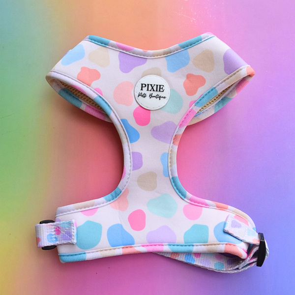 A pastel spot patterned dog harness with a white base and pink, blue, purple, and peach colours, featuring a round white brand patch that reads 'PIXIE Pets Boutique' on a colorful gradient background.